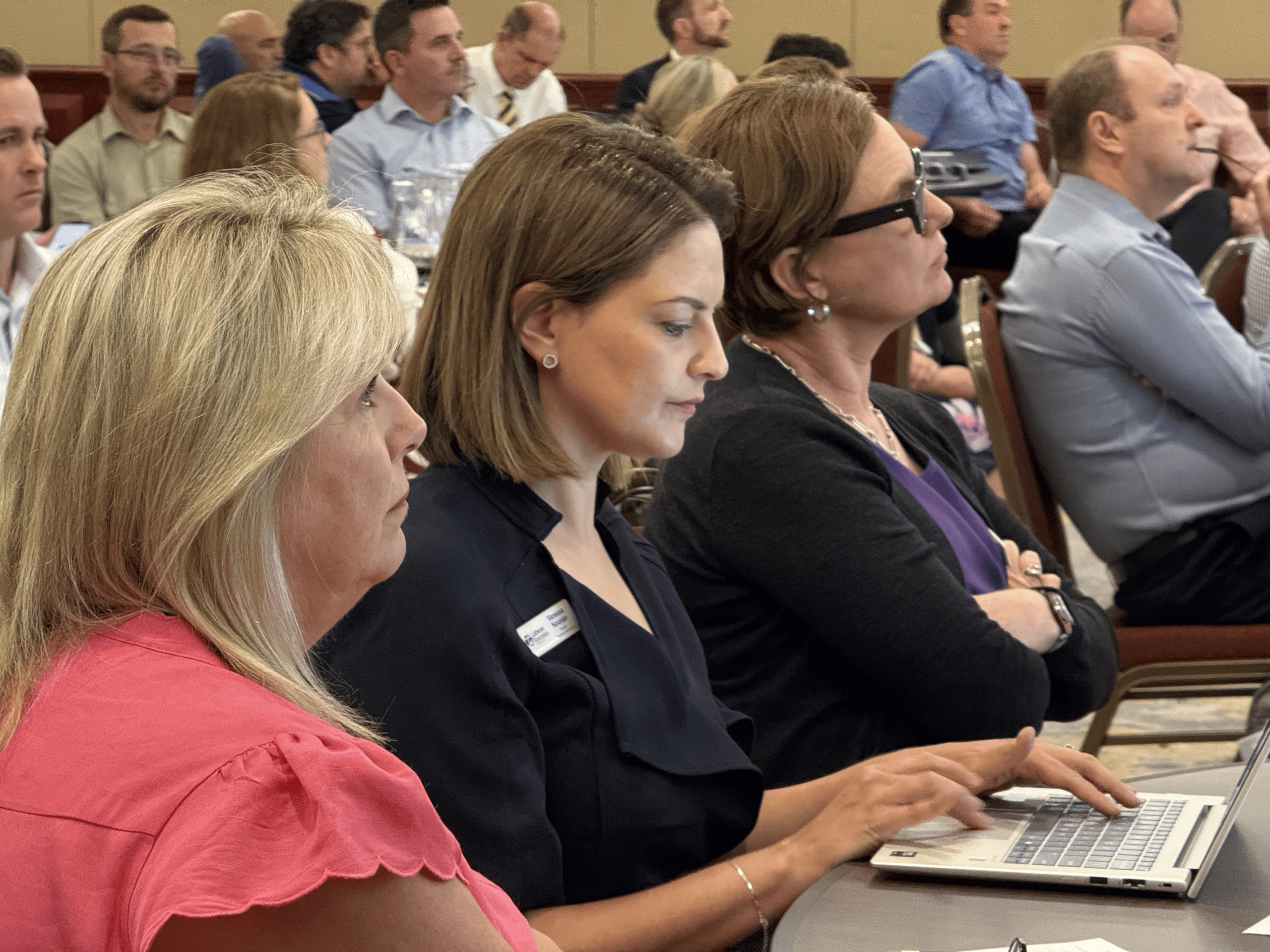 A group of people sit attentively in rows at a conference or seminar. In the foreground, three women focus on the presentation, with one typing on a laptop. Others listen and look forward.